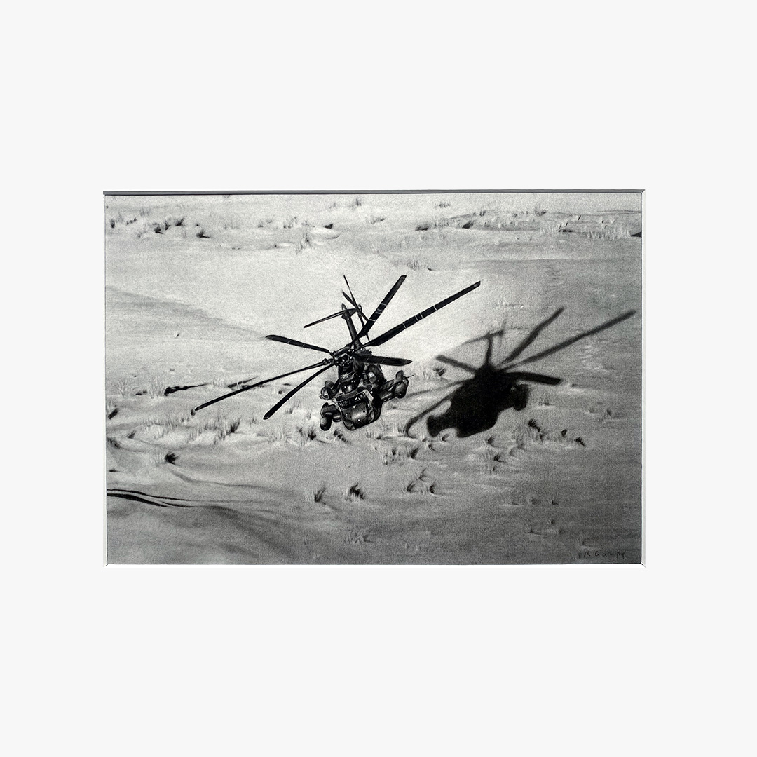 The image shows a black-and-white drawing of a helicopter flying over a desert with patches of dried grass. The helicopter's dark shadow is visible on the sand below.